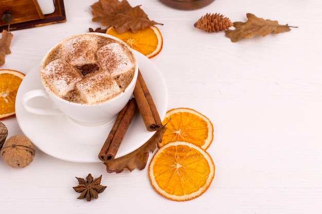 Cup of coffee with marshmallows and cocoa, leaves, dried oranges, spices, on a white background. Delicious hot autumn drink, morning mood. copyspace.