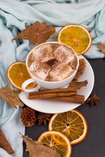 Cup coffee with marshmallows, cocoa, scarf, leaves, dried oranges, spices, on gray background. Delicious hot autumn drink, morning mood. copyspace.