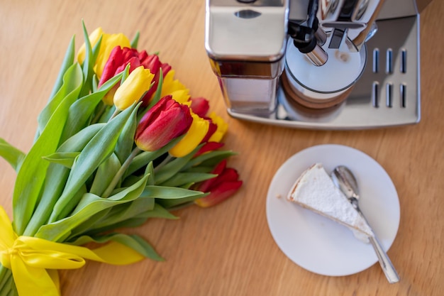 Cup of coffee with milk piece of cake and tulips flowers on wooden kitchen table Freshly brewed cappuccino espresso in coffee machine for breakfast Stylish home interior decor Flat lay top view