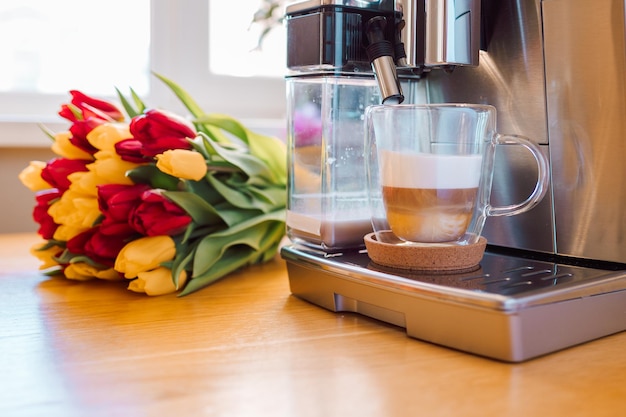 Cup of coffee with milk and tulips flowers on wooden kitchen table Freshly brewed cappuccino espresso in coffee machine for breakfast Stylish home interior decor