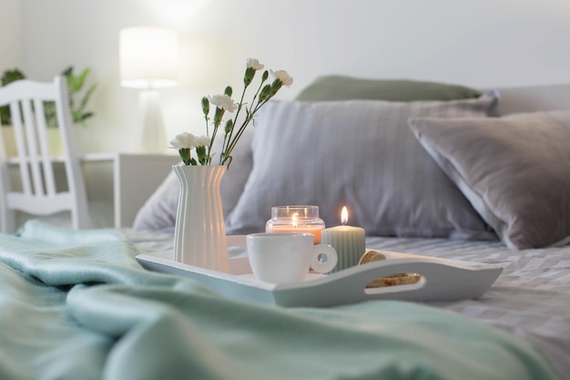 Cup of coffee on wooden tray on bed with flowers and candles