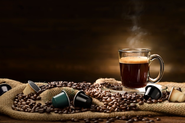 Cup glass of coffee with smoke and coffee beans and coffee capsules on burlap sack on old wooden table