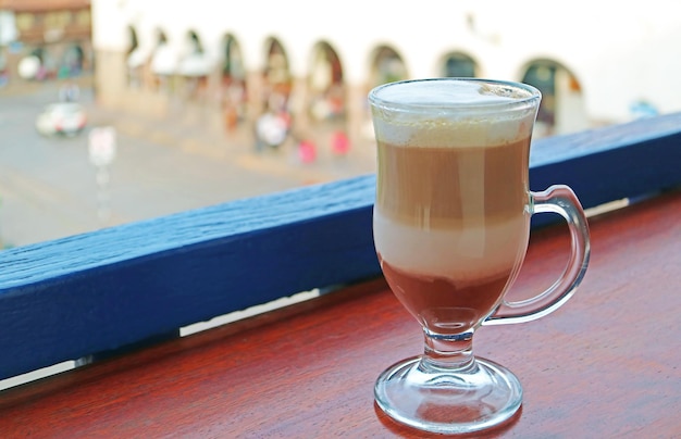 Cup of hot cappuccino coffee in a transparent cup with old city square of Cusco Peru in the backdrop