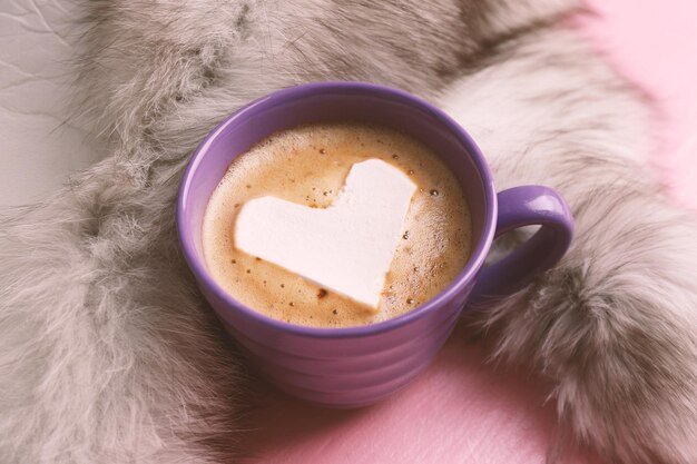 Cup of hot cappuccino with heart marshmallow and fur on pink background close up