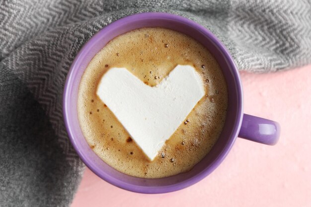 Cup of hot cappuccino with heart marshmallow and warm scarf on pink background close up