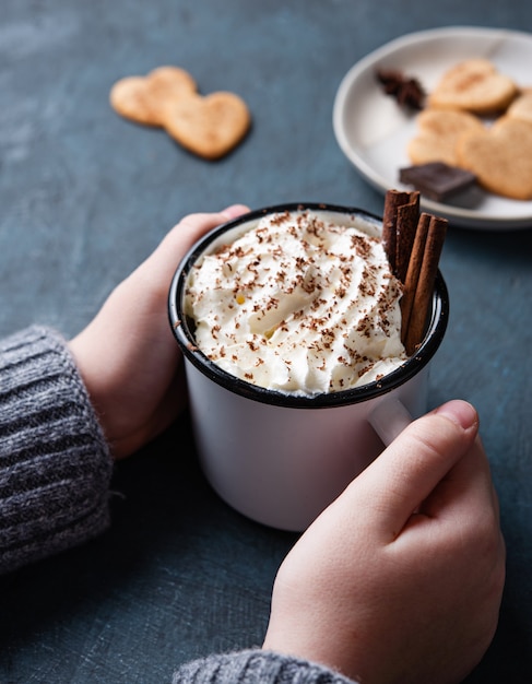 A cup of hot chocolate with cream and cinnamon  in  woman hand on a dark  table with homemade cookies. Top view and close up