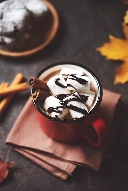 A cup of hot chocolate with marshmallows and cinnamon on a dark brown table with maple leaves and cookies Autumn concept of a warming drink Tint image