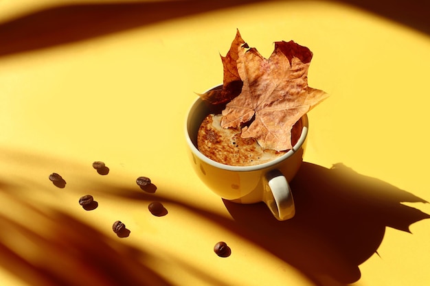 A cup of hot coffee will warm you on cold autumn days a yellow cup of coffee with a maple leaf in it scattered coffee beans on a yellow background shadows from objects