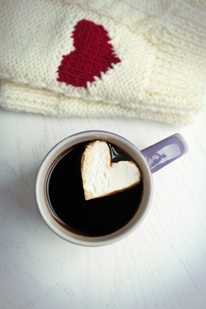 Cup of hot coffee with heart marshmallow and warm mittens on light wooden table, close up