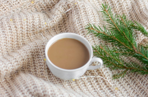 A Cup of hot coffee with milk on a warm blanket with a garland and a sprig of spruce.Christmas concept.spruce branch on a woolen blanket.