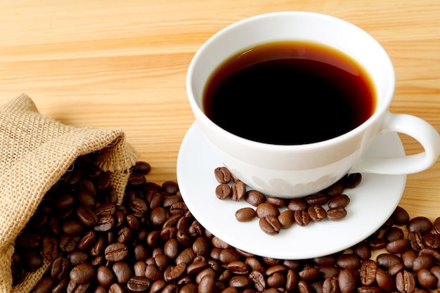 Cup of hot coffee with roasted coffee beans scattered from burlap bag on wooden table