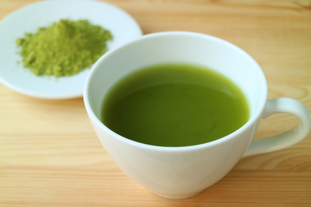 Cup of Hot Matcha Green Tea Served on Wooden Table with Blurry Plate of Matcha Tea Powder in Background