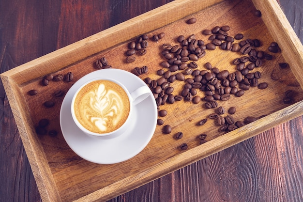 A Cup of latte art coffee and nearby coffee beans in a wooden box on a dark wooden table.