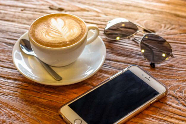 Cup of the latte art coffee with spoon and plate on the brown bark beautiful with warm light decorated with sunglasses and mobile phone