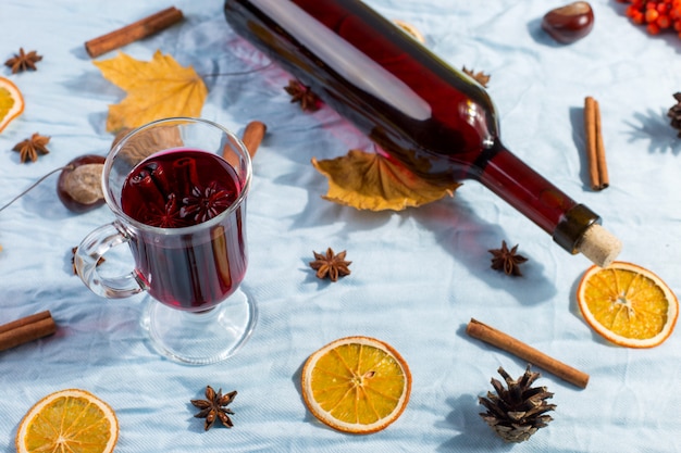 A cup of mulled wine with spices, bottle, dry leaves and oranges on table. Autumn mood, method to keep warm in the cold, copyspace, morning light.