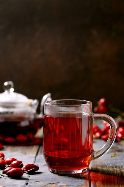 Cup of rose hip berries herbal tea and glass teapot standing on old wooden plank table with wild autumn berries around. Vitamin hot beverage.