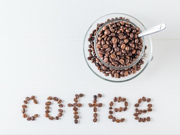 Cup and saucer filled with roasted coffee beans and coffee bean inscription on a white wooden table. 