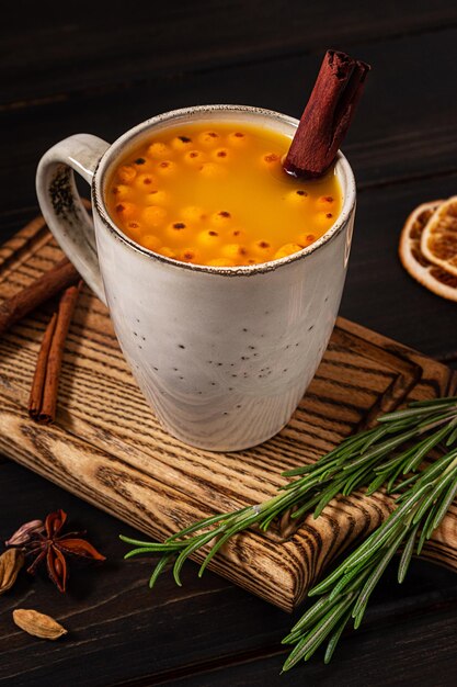 A cup of sea buckthorn hot drink, served with rosemary and cinnamon, on dark wooden background