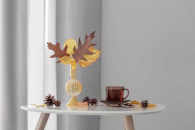 Cup of tea and autumn leaves in glass vase on background gray wall