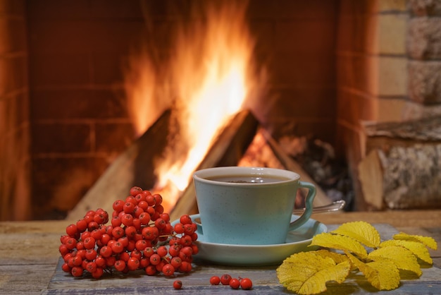 A cup of tea a bunch of mountain ash and yellow autumn leaves near a cozy fireplace