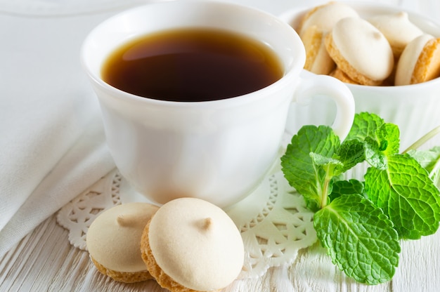 A cup of tea and small cookies with mint leaves on a white wooden table.