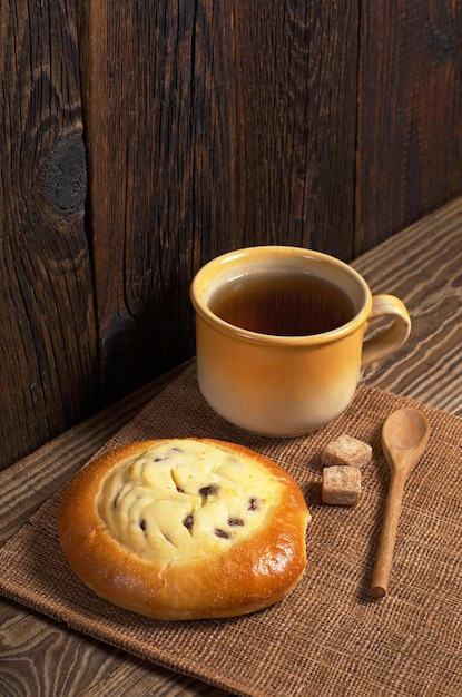 Cup of tea and tasty bun with cottage cheese and raisins for breakfast on dark wooden table