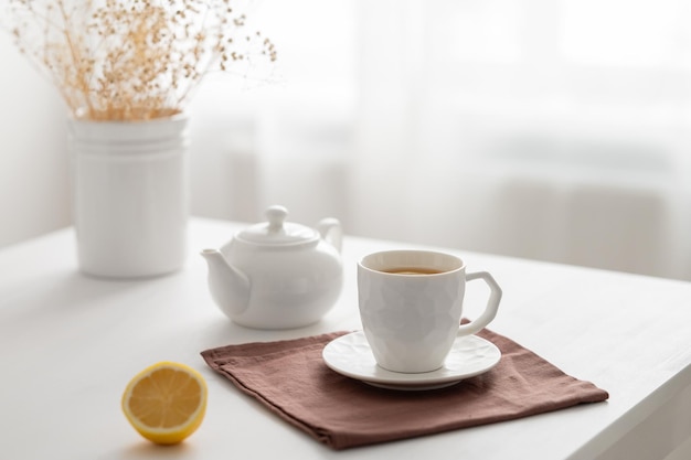 A cup of tea with lemon and a teapot on a white table with dry bouquet against the background of a kitchen window