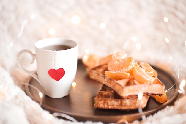 Cup of tea with red heart on it with waffles and oranges on wooden tray in bed Tasty breakfast Good morning