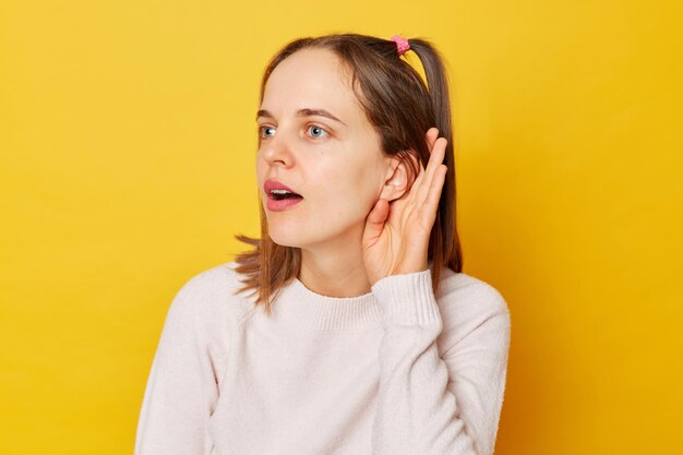 Photo curious attentive teenage girl in jumper with ponytails trying to overhear someone holding hand near ear tries to understand isolated over yellow background