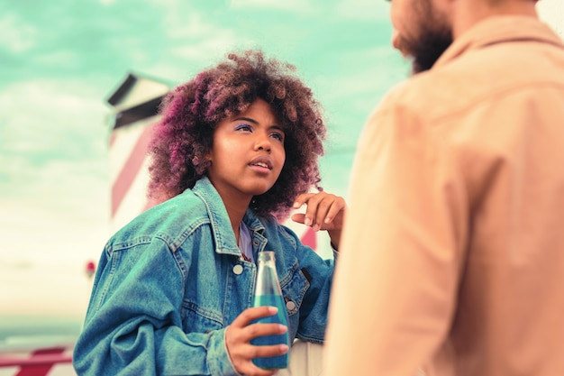 Curious girl. Attentive thoughtful young girl sitting with a glass bottle in her hand and looking attentively at the young man while listening to him