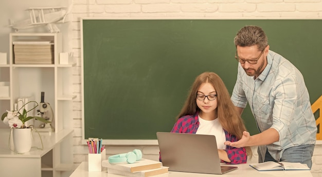 Curious kid and dad sitting in classroom with computer at blackboard back to school