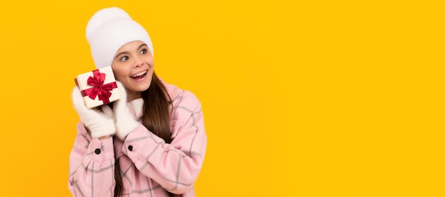 Curious kid in hat and mittens hold gift box on yellow background new year Banner of christmas child girl studio kid winter portrait with copy space