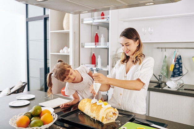 Curious little girl looking at her mother decorating swiss roll with orange slices and whipped cream