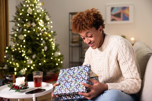 Curious man sitting on couch near christmas tree smiling handsome guy in white sweater