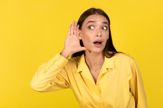 Curious millennial woman listening secrets eavesdropping posing over yellow background