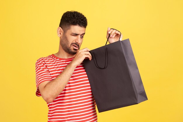 Photo curious nosy man with beard striped t-shirt peeking into black paper shopping bag, unpacking gift, trying to guess what is inside, preparing surprise. indoor studio shot isolated on yellow background