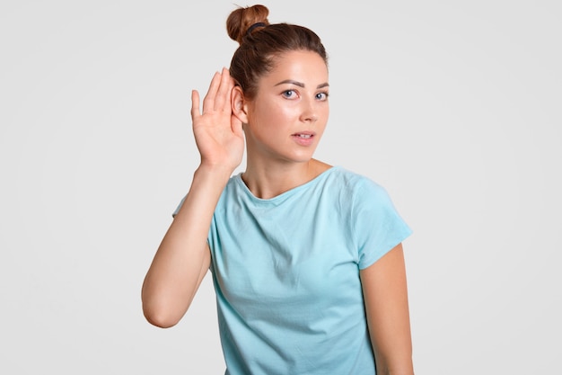 Curious woman puts hand near ear, tries to eavesdrop or overhear secret information, likes gossiping, dressed in casual blue t shirt, isolated over white. Speak loudly to hear better you