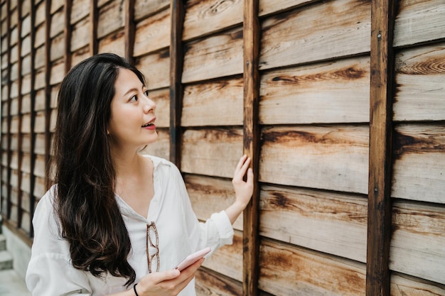 curious young asian japanese lady tourist walking along wooden wall and holding cellphone with online guide book sightseeing in monument. joyful smiling girl traveler with mobile phone relax outdoor