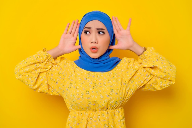 Curious young beautiful Asian Muslim woman holding hands near ears trying to listen secret isolated over yellow background