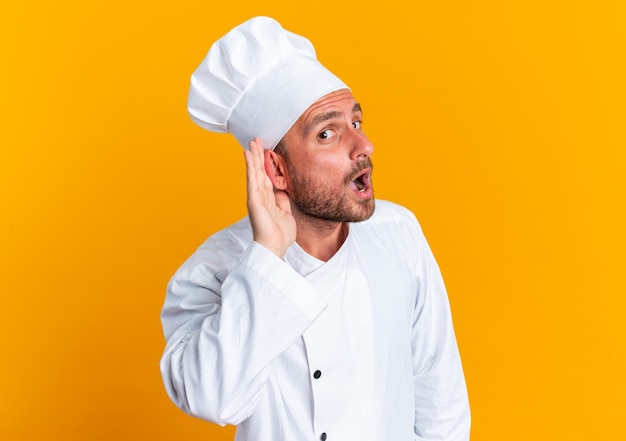 Curious young caucasian male cook in chef uniform and cap looking at camera doing i can't hear you gesture isolated on orange wall