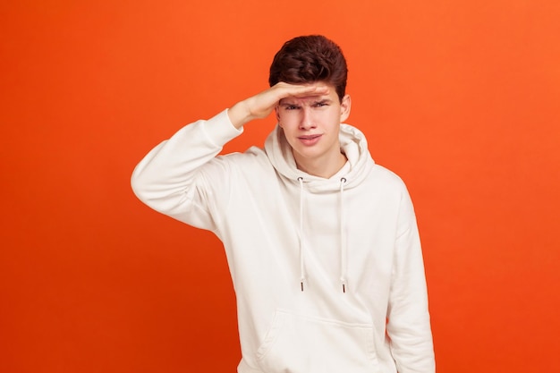 Curious young man in casual hoodie looking far away with hand over head, student looking for job, future prospects. Indoor studio shot isolated on orange background