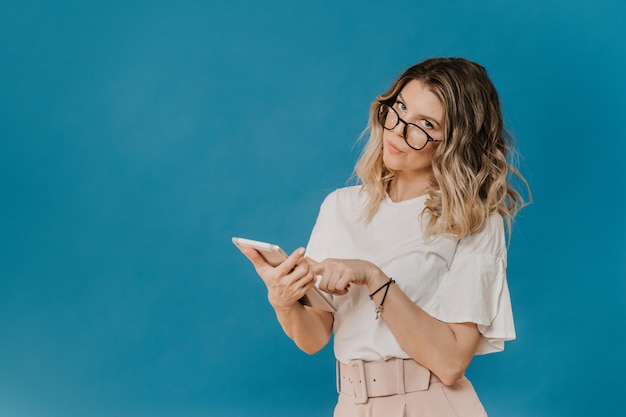 Curly blonde in glasses dressed a light t shirt pink pants with a belt upset by the news she read on her tablet, points at the screen, over blue background with empty space. Networking people concept