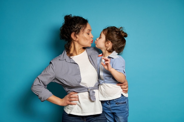 Curly brunette woman and her cute baby girl kissing while looking to each other on blue surface with copy space