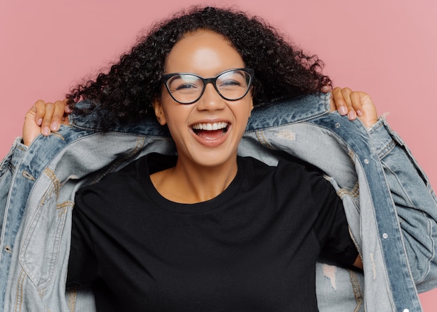 Curly cheerful woman tries on new denim jacket, wears optical glasses, smiles broadly, has perfect mood