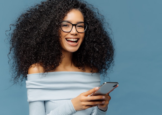 Curly cheerful woman with pleased expression, holds mobile phone, checks email box, wears optical glasses and jumper, enjoys using modern technologies