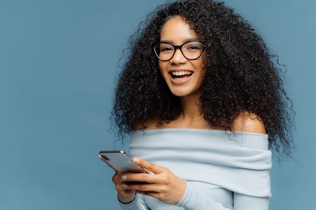 Curly cheerful woman with pleased expression, holds mobile phone, checks email box