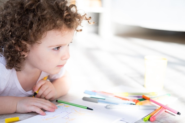 Curly child girl drawing with colored markers lying on floor in living room at home