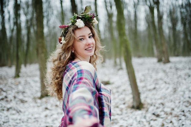 Curly cute blonde girl with wreath in checkered plaid at snowy forest in winter day.