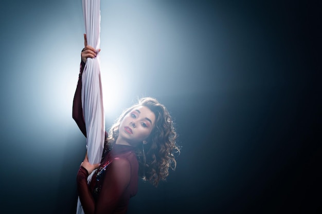 Curly female gymnast performs on a black background with rays circus artist on white aerial silks