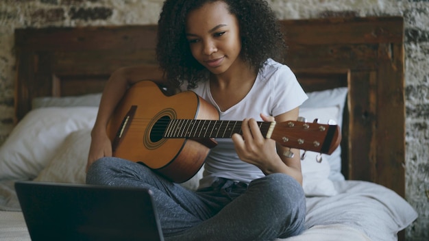 Curly girl concentrating learning to play guitar using laptop computer sitting on bed at home